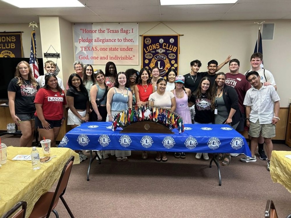 Kilgore Lions Club members and American Exchange Students gathered around a table with a blue Lions Club tablecloth and a flag display